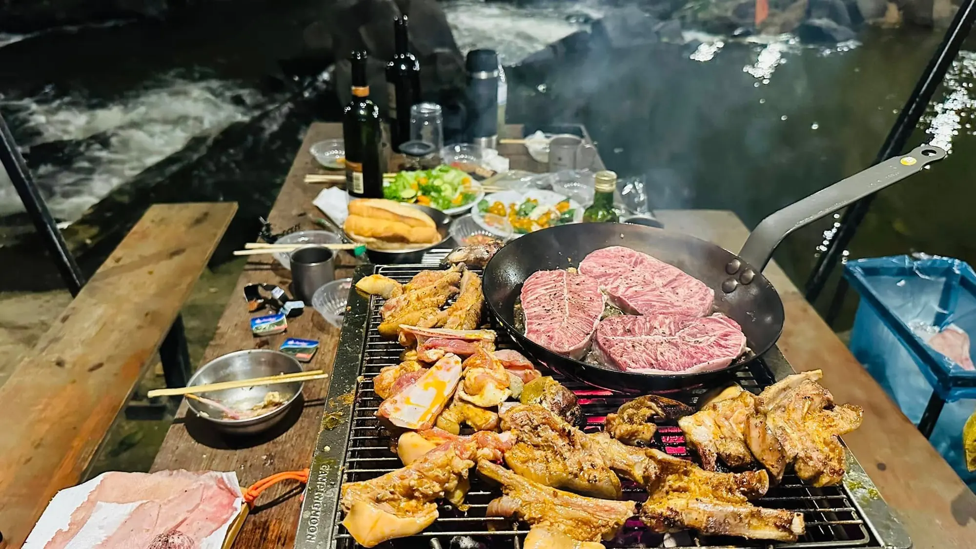 Bếp nướng BBQ với thịt, xúc xích và chảo thịt bò bên suối buổi tối