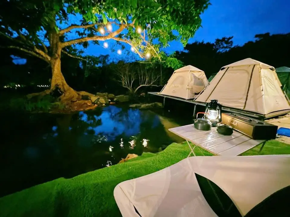 Hai lều dựng sẵn bên vũng suối lúc chạng vạng, cây lớn giăng đèn dây, bàn có đèn bão và ấm nước
